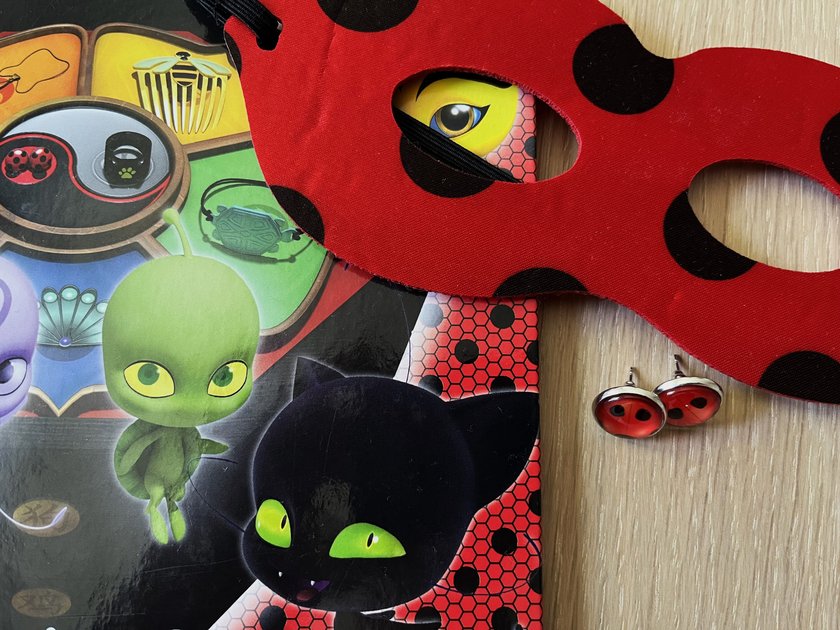 Miraculous-Spielzeug: Marienkäfer-Maske und Marienkäfer-Ohringen liegen neben Miraculous-Buch
